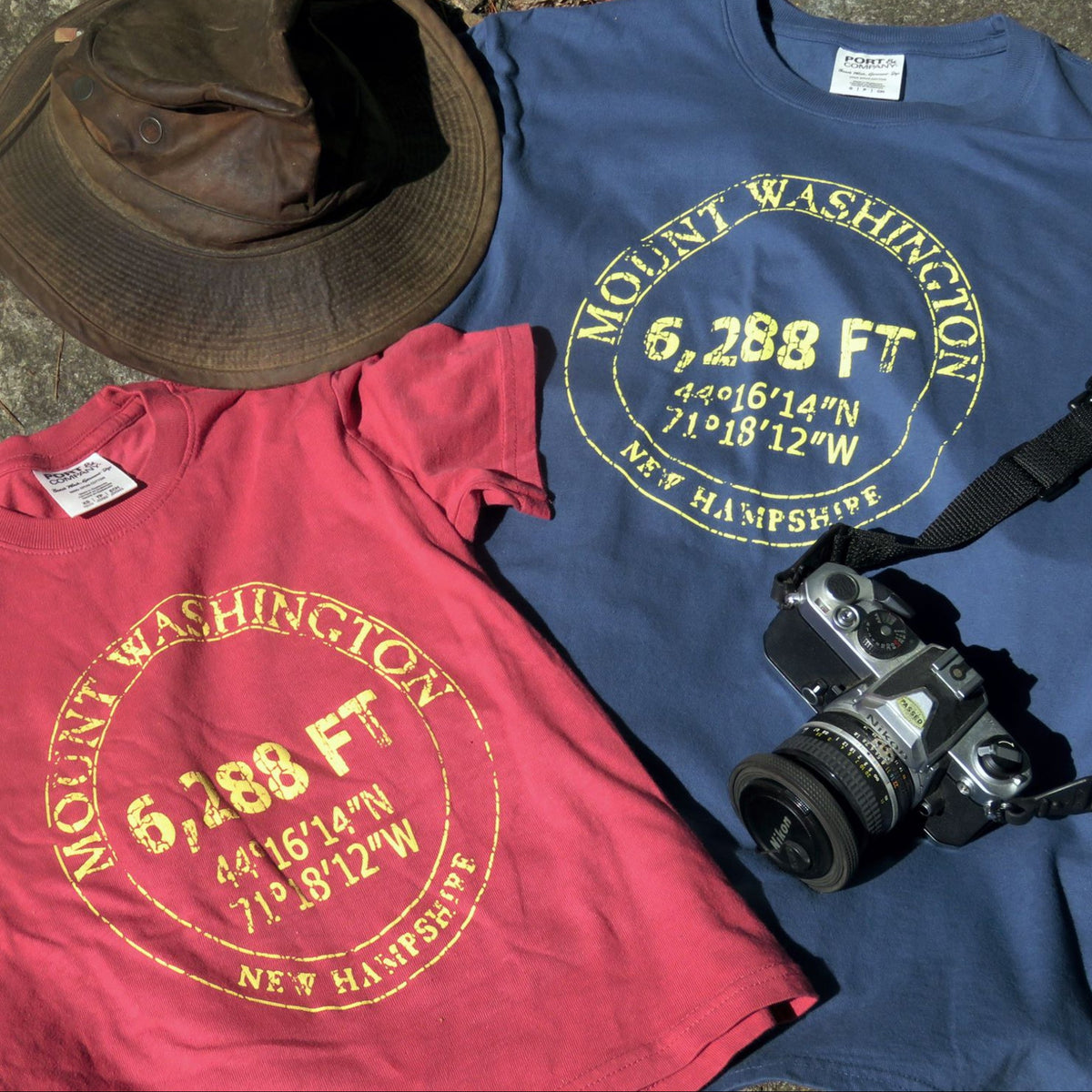 Two t-shirts with Mount Washington design, one red and one blue, with a camera and hat on top.