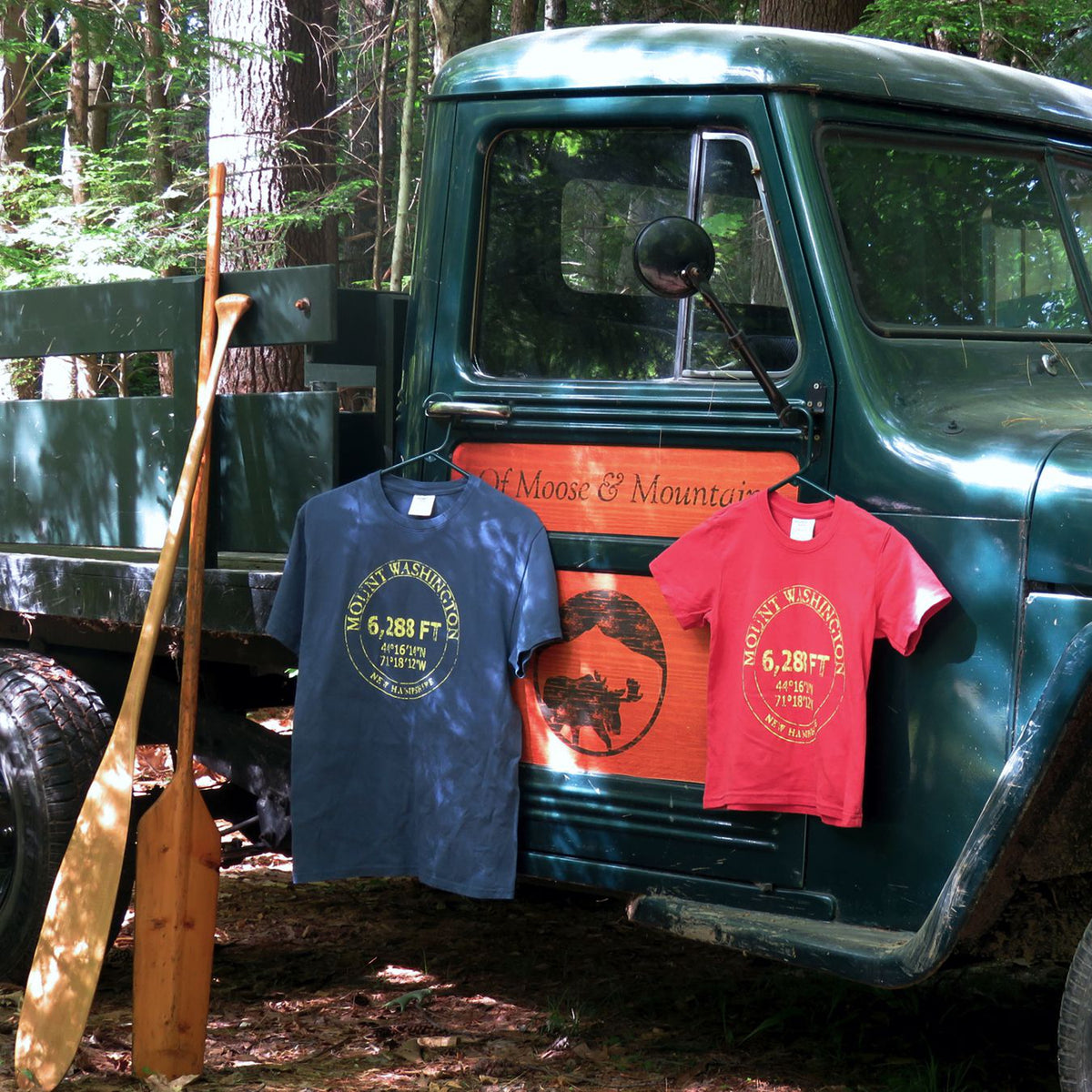 Two t-shirts hanging on a vintage truck in a forest setting with a paddle leaning against the truck.
