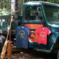 Two t-shirts hanging on a vintage truck in a forest setting with a paddle leaning against the truck.