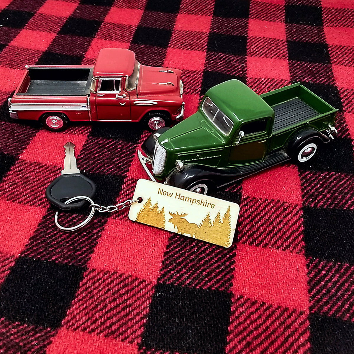 Two toy trucks, one red and one green, on a red and black checkered fabric with a keychain and 'New Hampshire' tag.