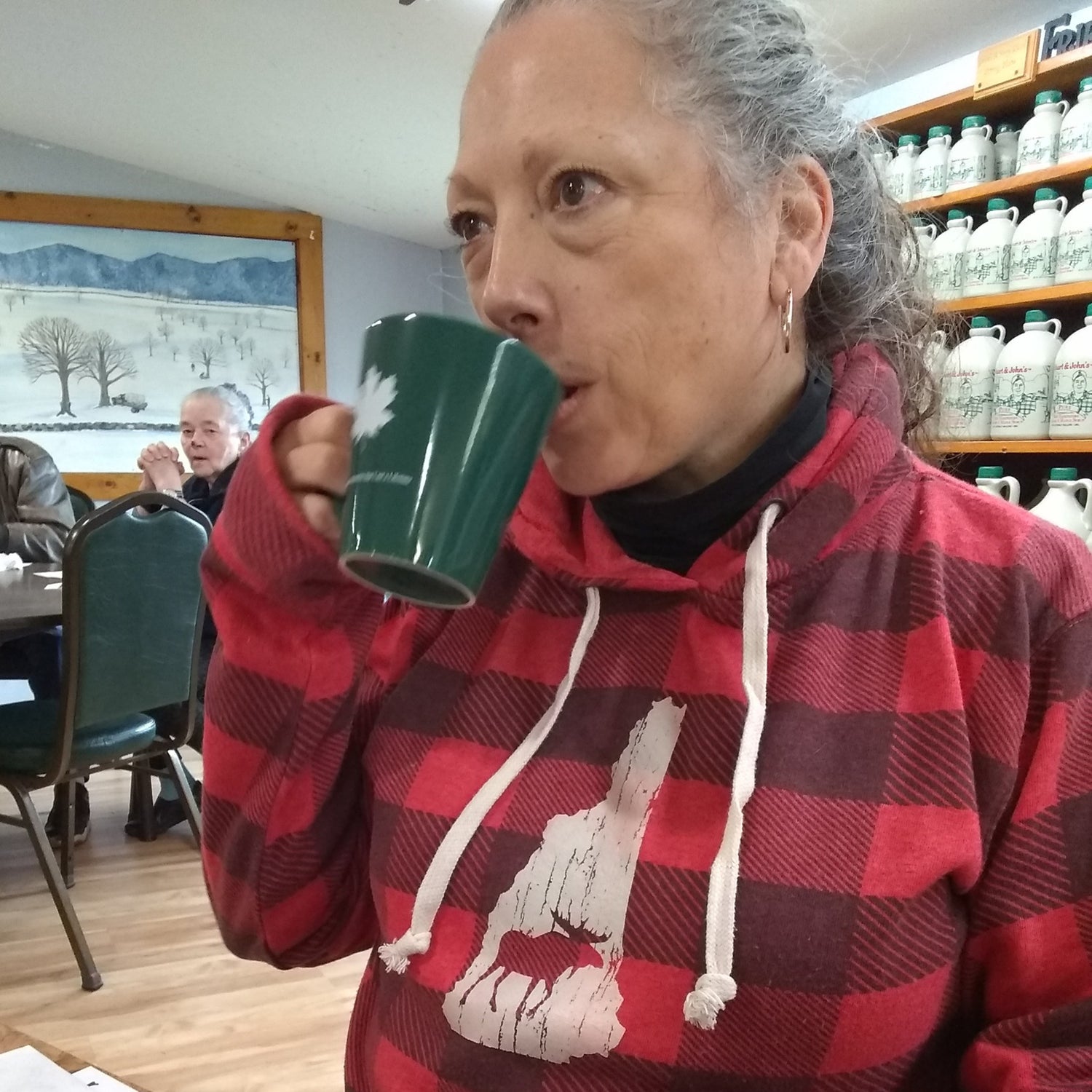 Woman in a red plaid hoodie drinking from a green mug in a room with shelves and another person in the background.