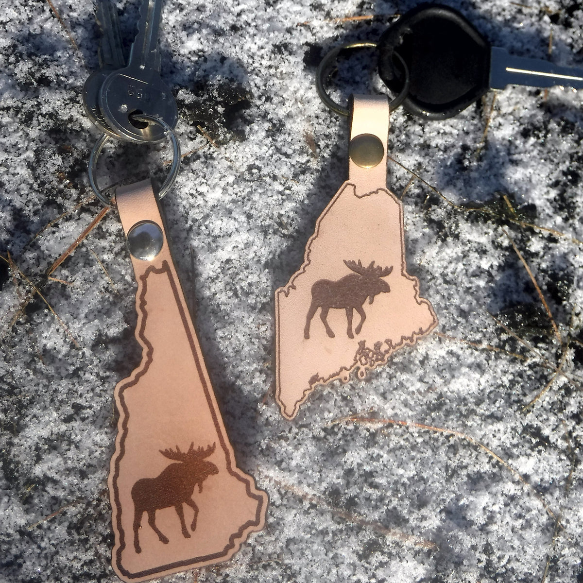 Two leather keychains shaped like the states of New Hampshire and Maine with moose designs on a textured surface.