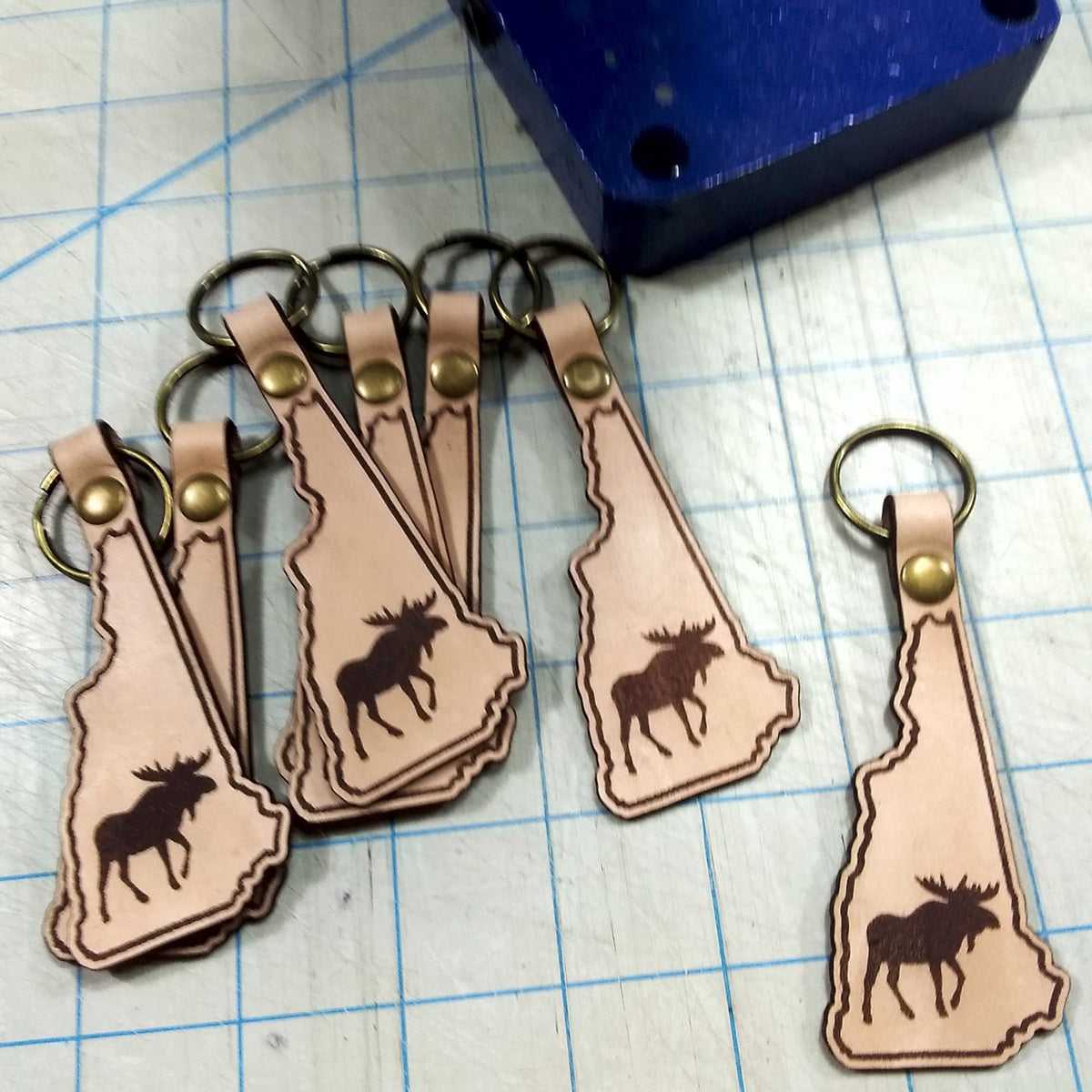 Leather keychains shaped like New Hampshire with moose silhouette on a grid surface