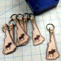 Leather keychains shaped like New Hampshire with moose silhouette on a grid surface
