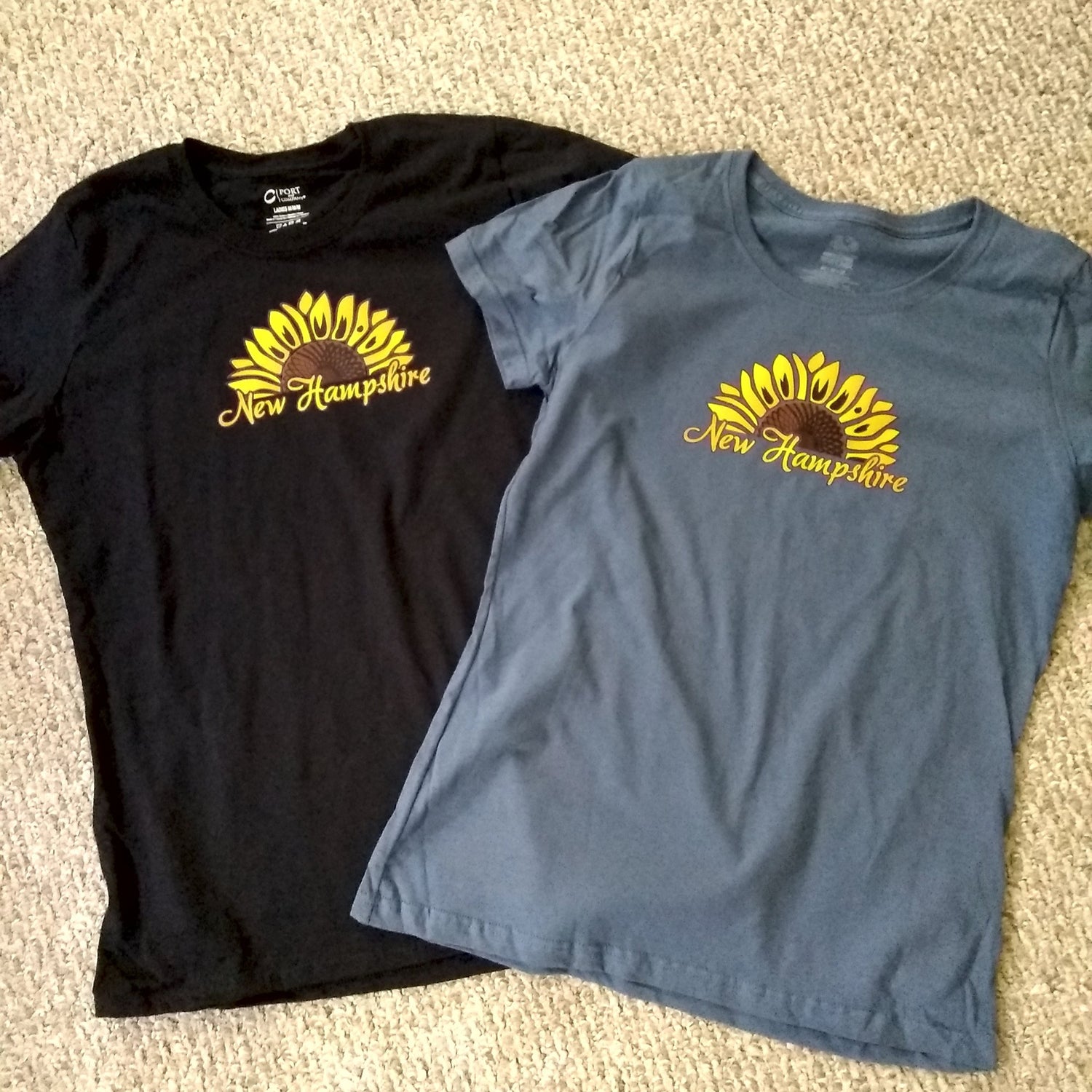 Black and blue t-shirts with sunflower design and 'New Hampshire' text on a beige carpet.