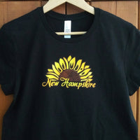 Black t-shirt with yellow sunflower design and 'New Hampshire' text on a wooden background