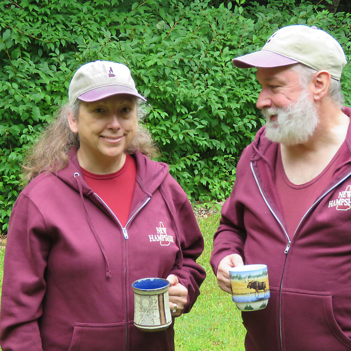 Two people wearing maroon hoodies and caps with visible branding, holding mugs outdoors.