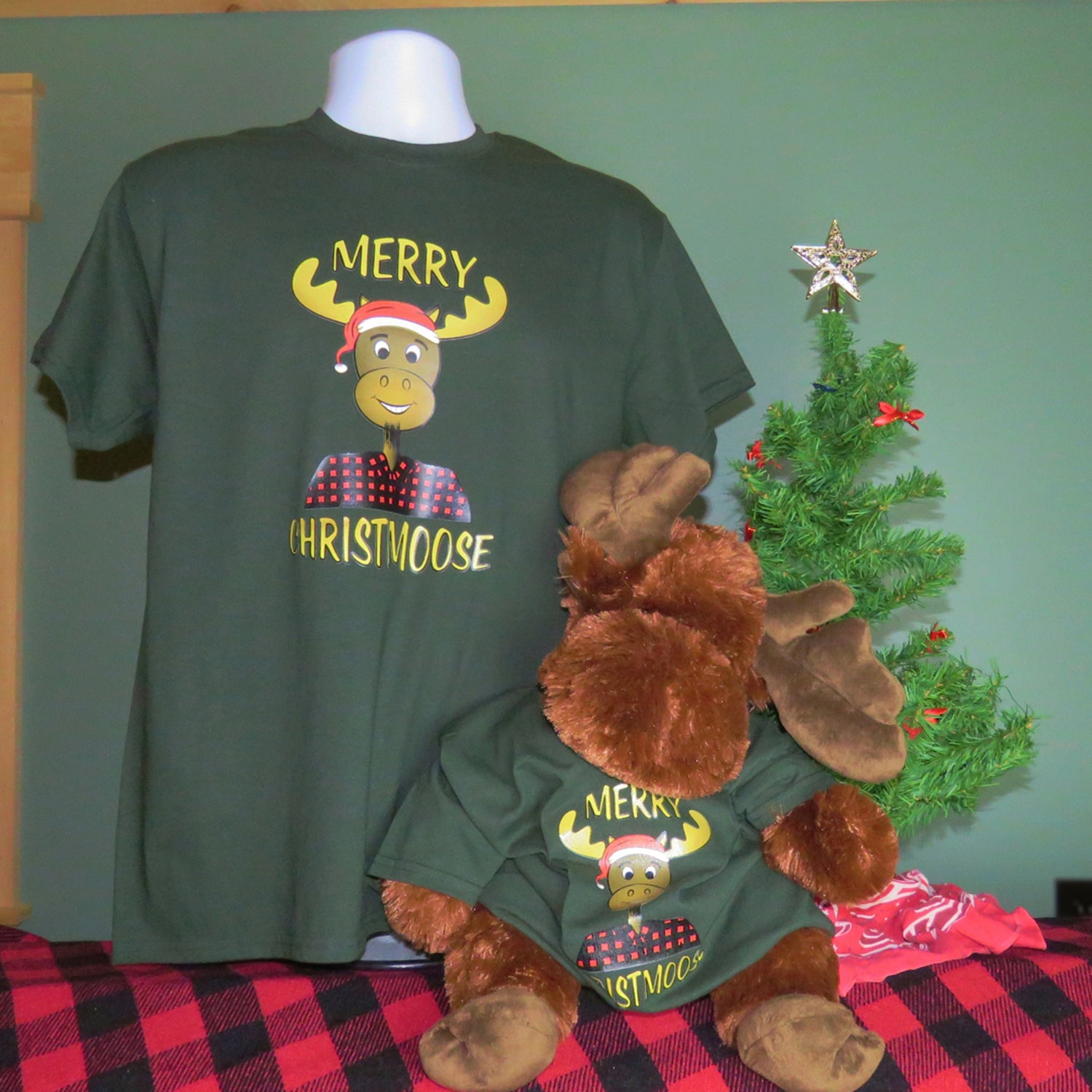 Green t-shirt with 'Merry Christmoose' design and matching plush moose toy on a checkered tablecloth.