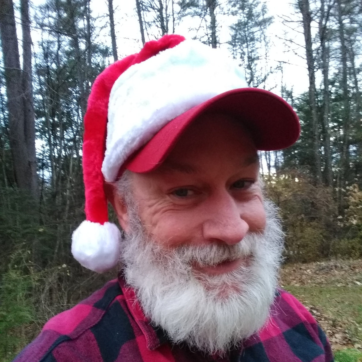 Man wearing a red and white Santa hat with baseball cap bill outdoors with trees in the background