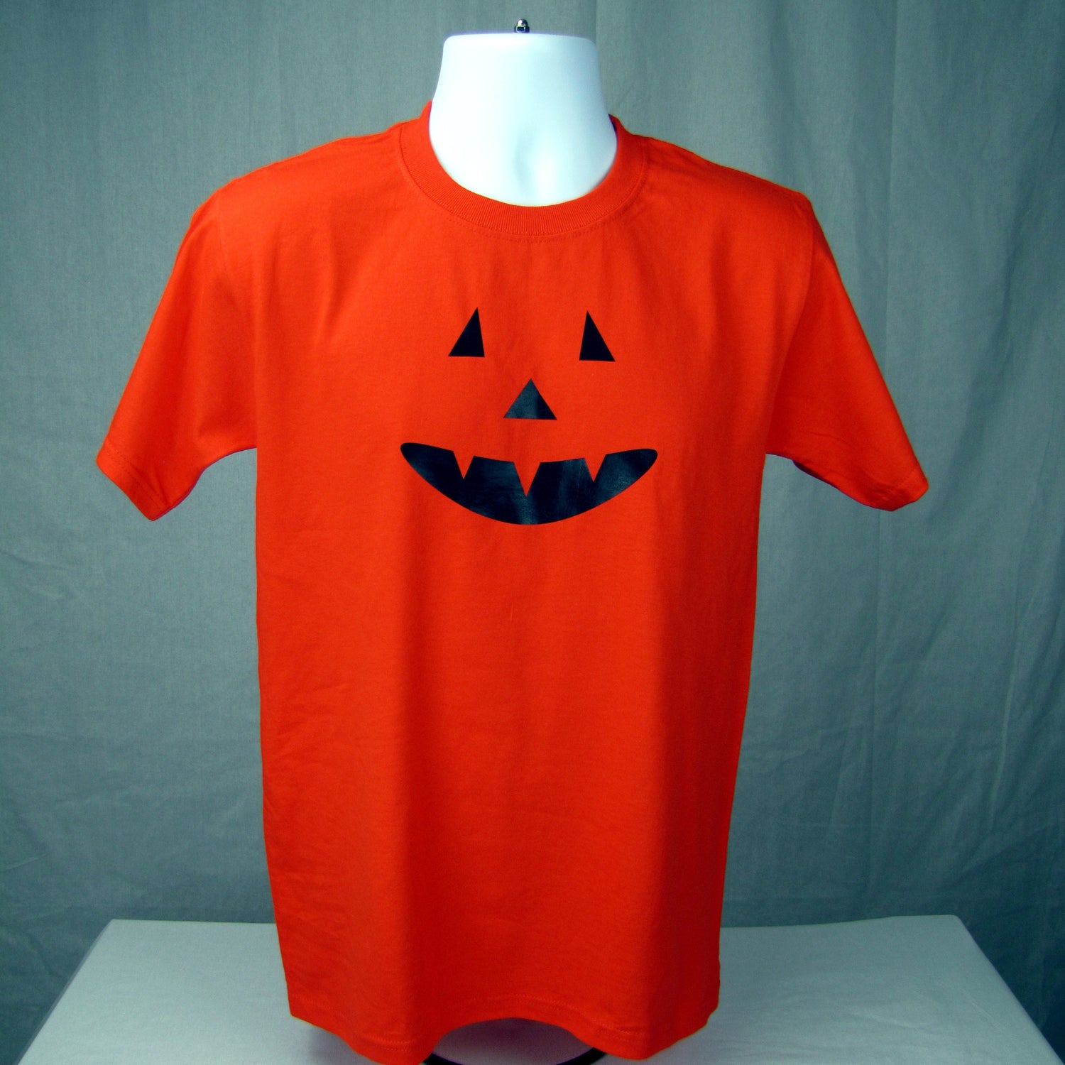 Orange t-shirt with black jack-o'-lantern face design on a mannequin against a green background