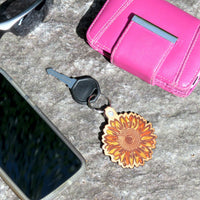 Keychain with sunflower design on a concrete surface next to a pink wallet and phone.