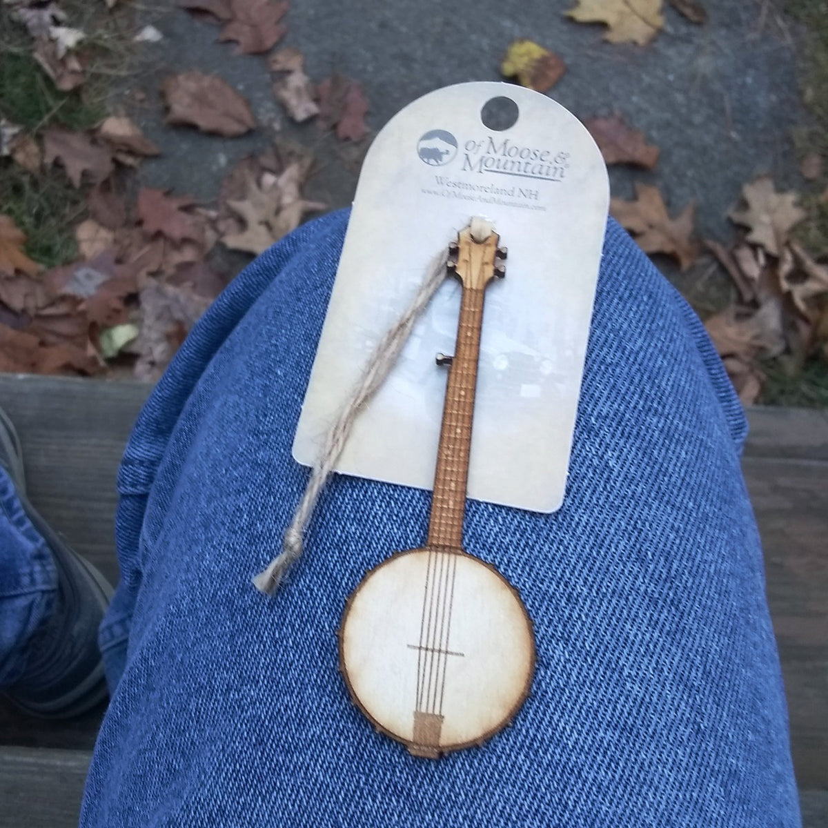 Small wooden banjo ornament on a blue denim surface with a brand tag in the background.