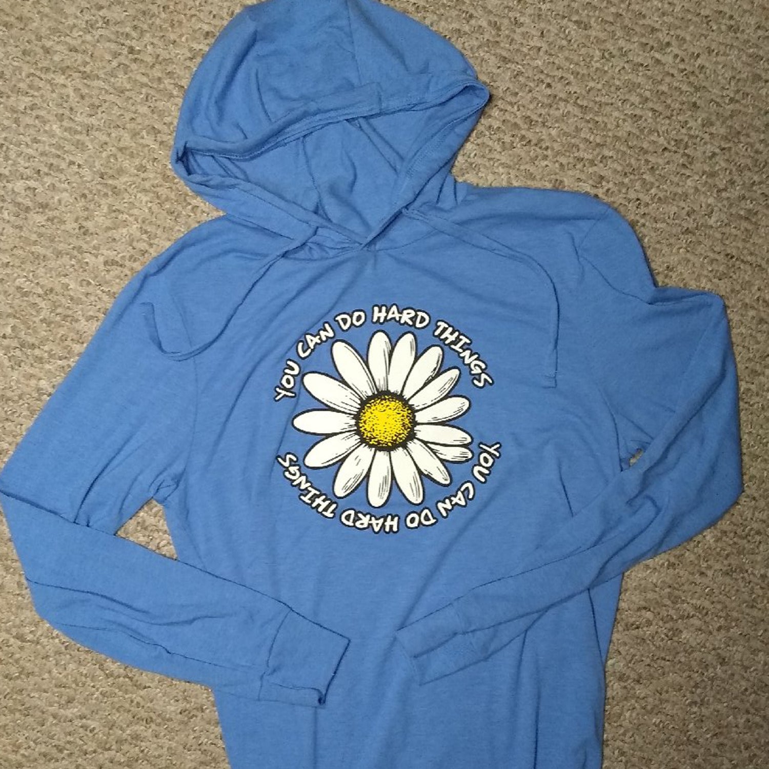 Blue hooded t-shirt with a daisy and motivational text on a beige carpet