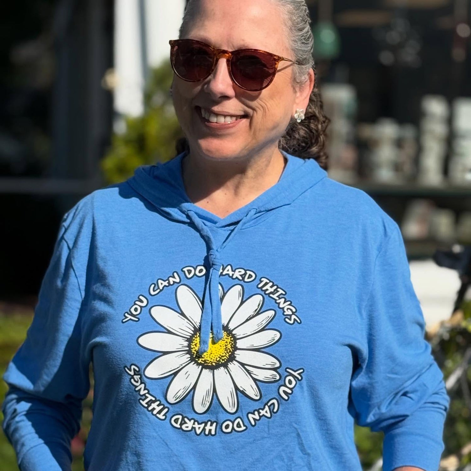 Person wearing a blue hooded t-shirt with a daisy design and uplifting text outdoors