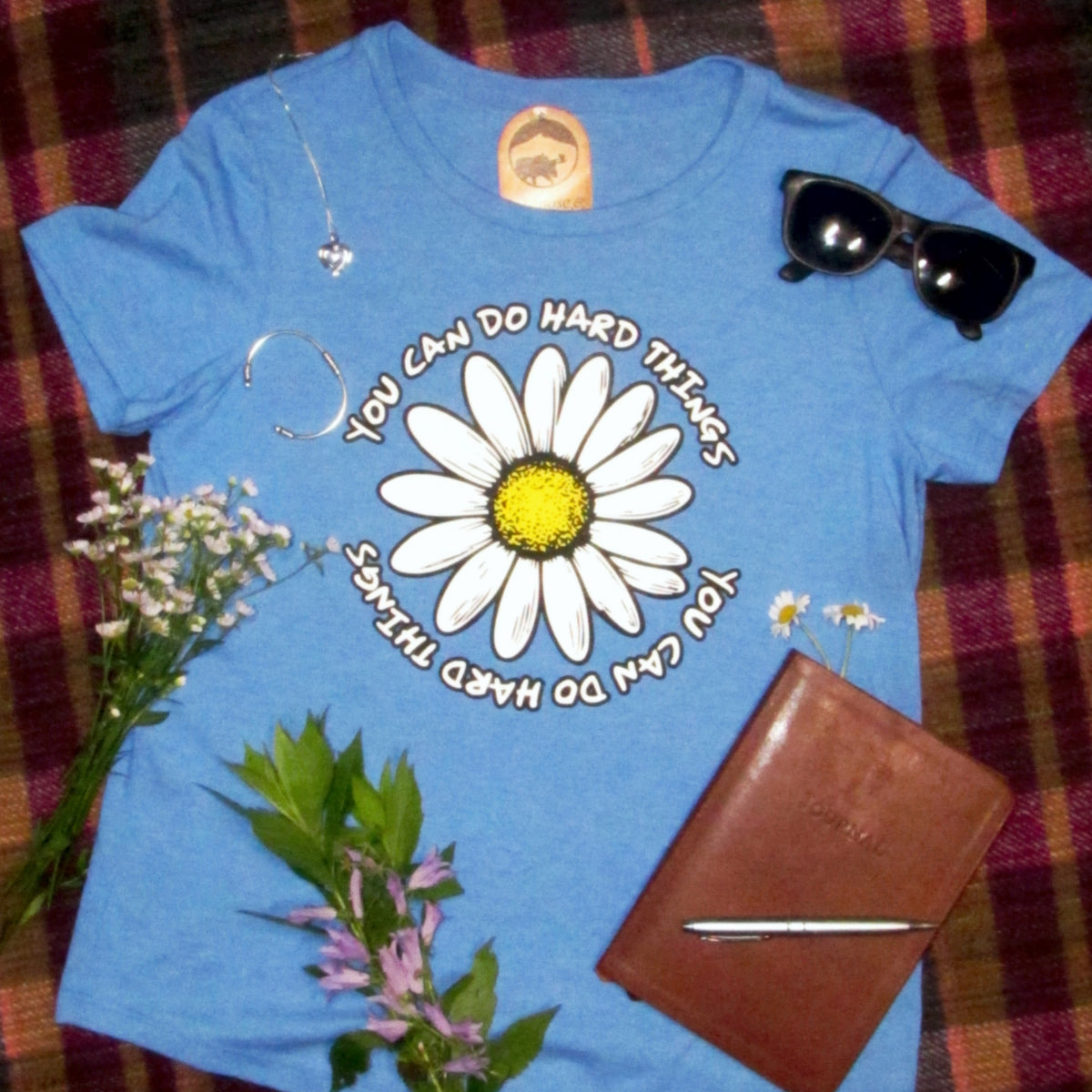 Blue t-shirt with a daisy graphic and text on a plaid background, surrounded by flowers, sunglasses, and a notebook.