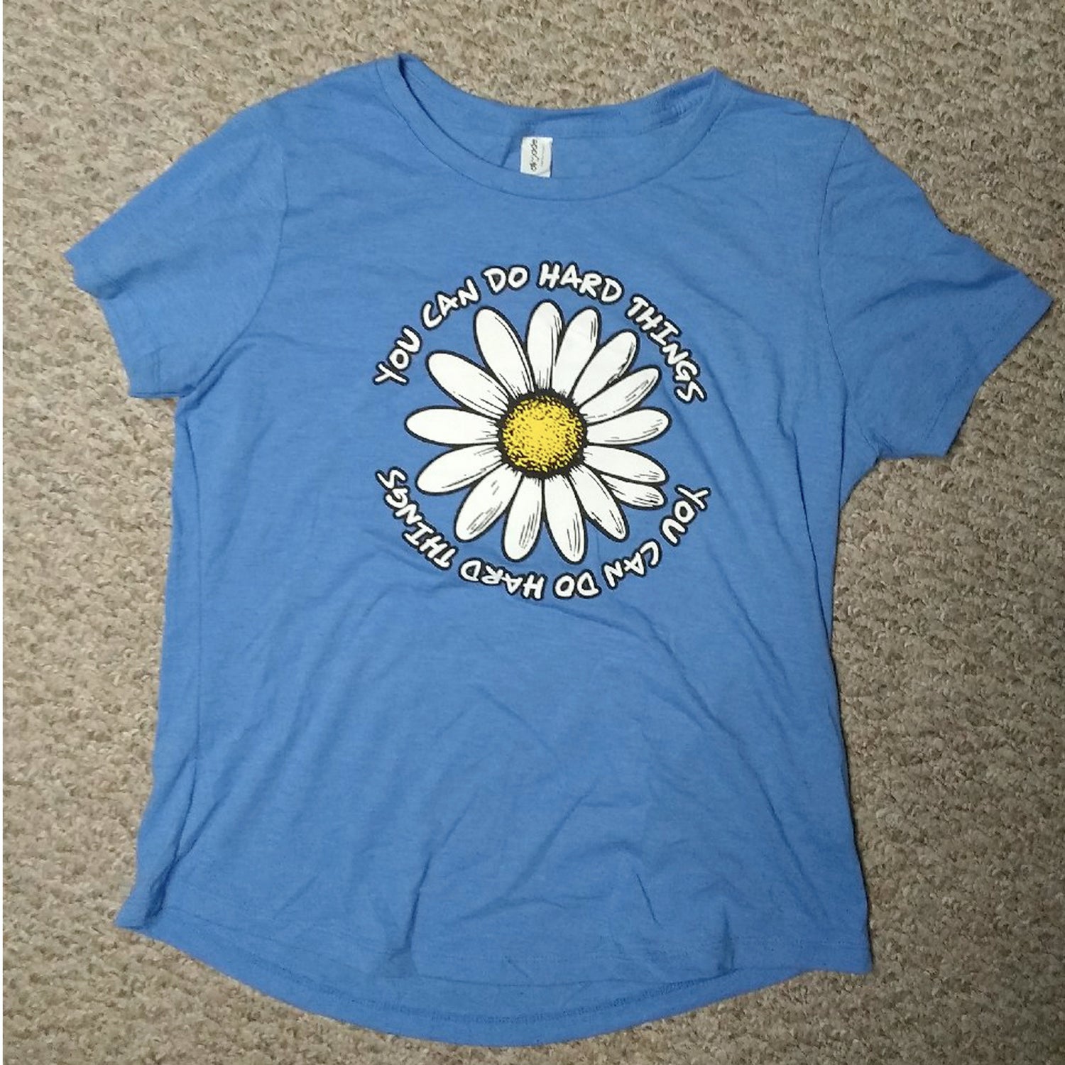 Blue t-shirt with a daisy graphic and text on a beige carpet