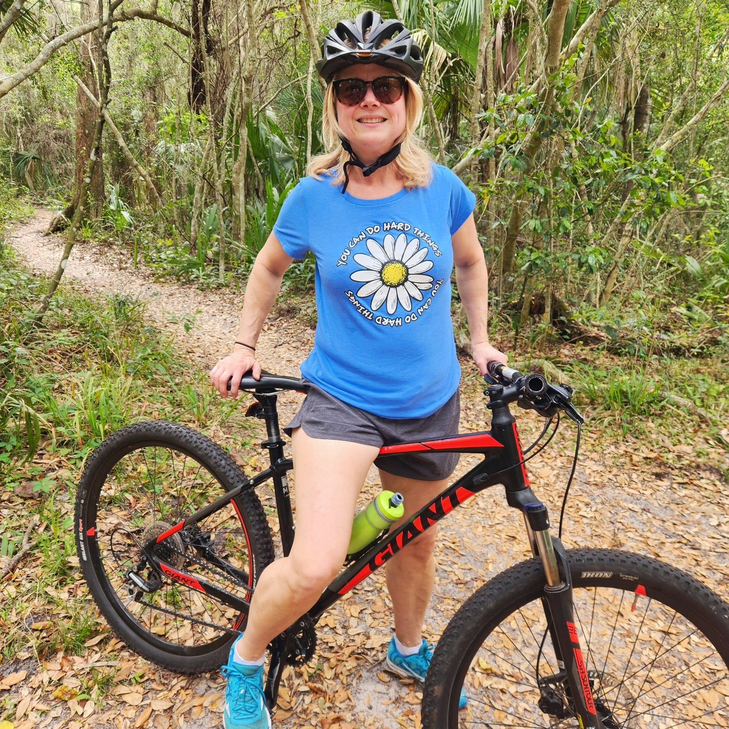 Woman with a mountain bike in a forest setting wearing a blue shirt with a daisy and inspirational text printed on it.