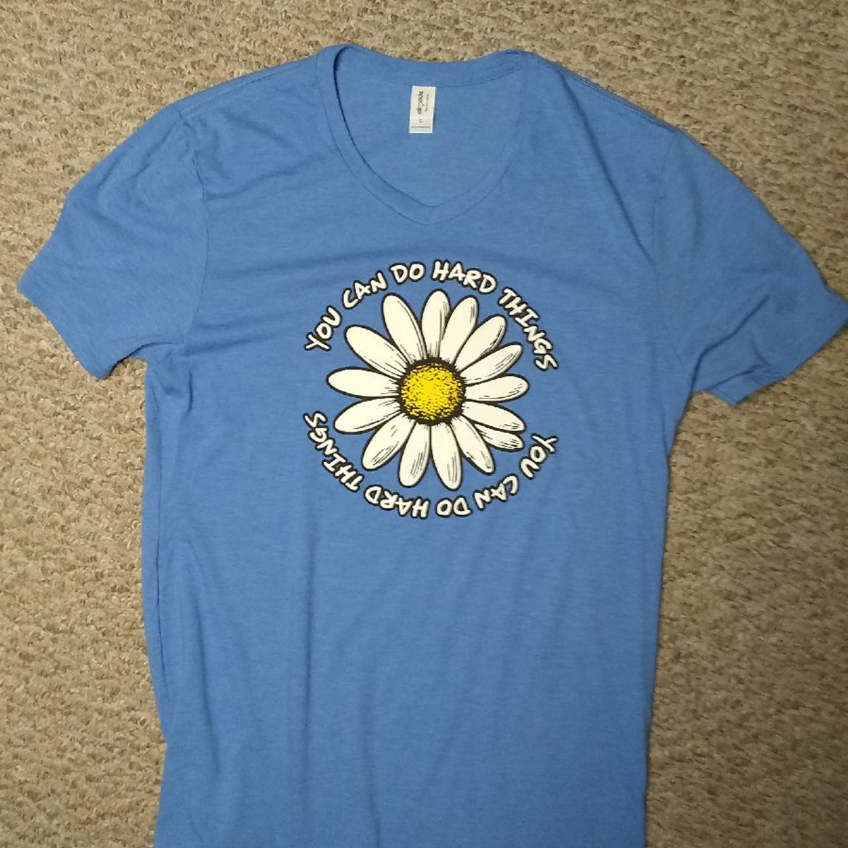 Blue t-shirt with a daisy graphic and text on a beige carpet background