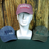 Three baseball caps with 'MAINE' text on a mannequin head and wooden background