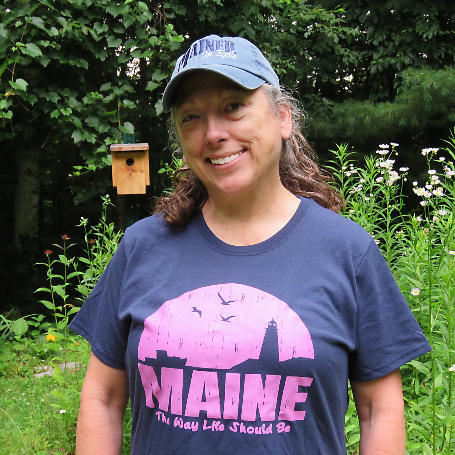Woman wearing a blue 'MAINE' t-shirt and cap in a garden setting
