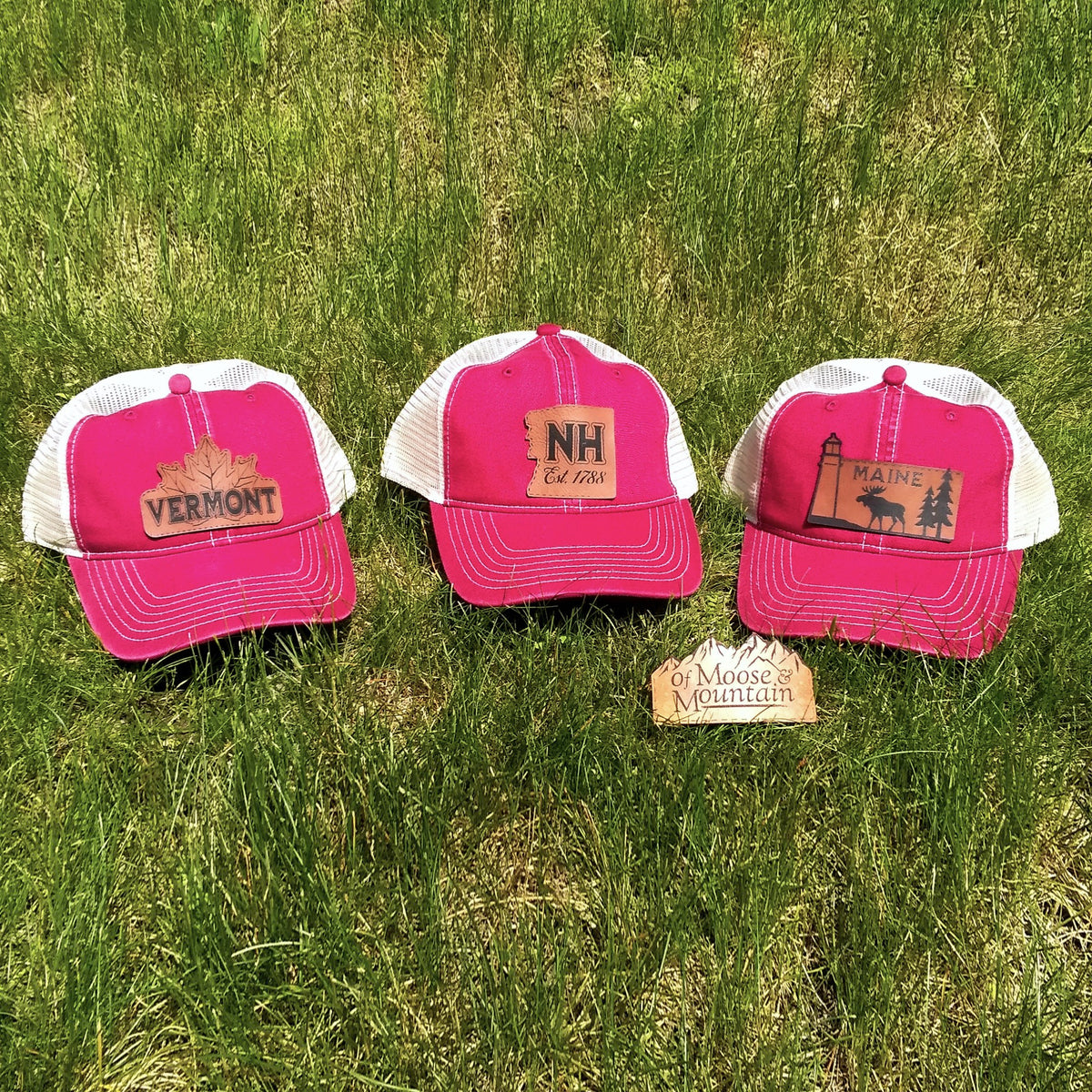 Three red and white trucker hats with state designs on a grassy background